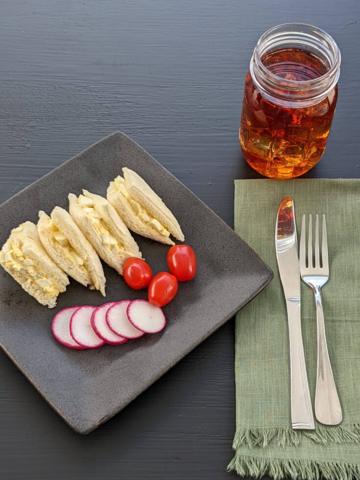 Egg salad sandwich on a plate with radish slices, grape tomatoes, a glass of iced tea, a green napkin, a knife and a fork on a black background
